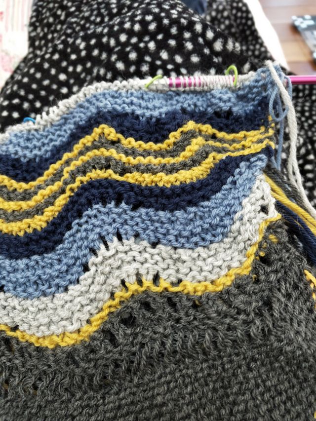 Image of my hansel hap. A garter stitch shawk. Visible are a lace (feather and fan) in coloured stripes (dk grey, mustard, light grey, light blue and dark blue)